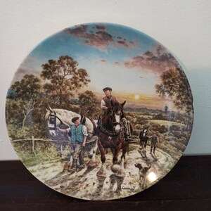 May include: A vintage ceramic plate with a colorful painting of a horse-drawn carriage on a country road. The scene includes two men, a dog, and a windmill in the distance. The plate has a blue and white border.