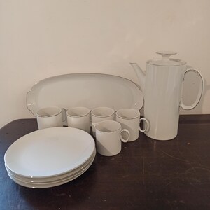 May include: A white porcelain tea set with a silver trim. The set includes a teapot, creamer, sugar bowl, and four cups and saucers. The set is displayed on a wooden table.