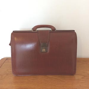Pode incluir: Uma pasta de couro castanho vintage com uma pega e um fecho de metal. A pasta tem uma forma retangular e um aspeto ligeiramente gasto. A pega é castanho mais escuro e o fecho de metal é de cor latão. Cerca de 40 cm de largura.