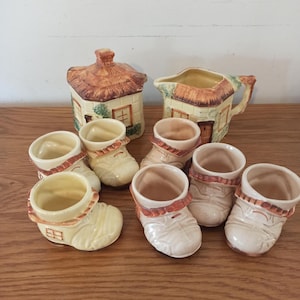 May include: A collection of ceramic tableware featuring a cottage theme. Includes a sugar bowl, creamer, and several boot-shaped egg cups. The pieces are primarily cream-colored with brown and orange accents, and a thatched roof design.