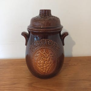 Op de afbeelding: Een bruine keramische Rumtopf-pot met een deksel, handvatten en een druivenontwerp. De pot heeft verticale lijnen en het woord "RUMTOPF" is in reliëf op de voorkant. De pot staat op een houten oppervlak.