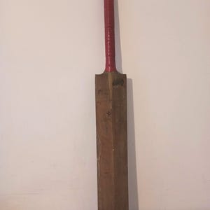 May include: A vintage cricket bat made of wood, with a red grip handle. The bat is brown with a rounded end and is leaning against a white wall. The handle is wrapped in red tape.