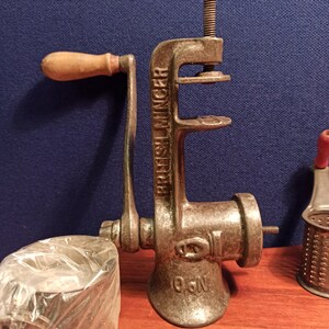 May include: A vintage cast iron hand-crank meat grinder with a wooden handle. The grinder has a label that reads "British Mincer" and a number "06N".