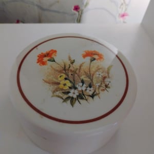 Puede incluir: Una caja de cerámica blanca con un borde marrón y un diseño floral con flores naranjas, amarillas y blancas.