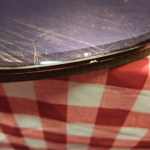 May include: Close-up of the edge of a round, dark purple table with a clear epoxy resin finish. The table is sitting on a red and white checkered tablecloth.