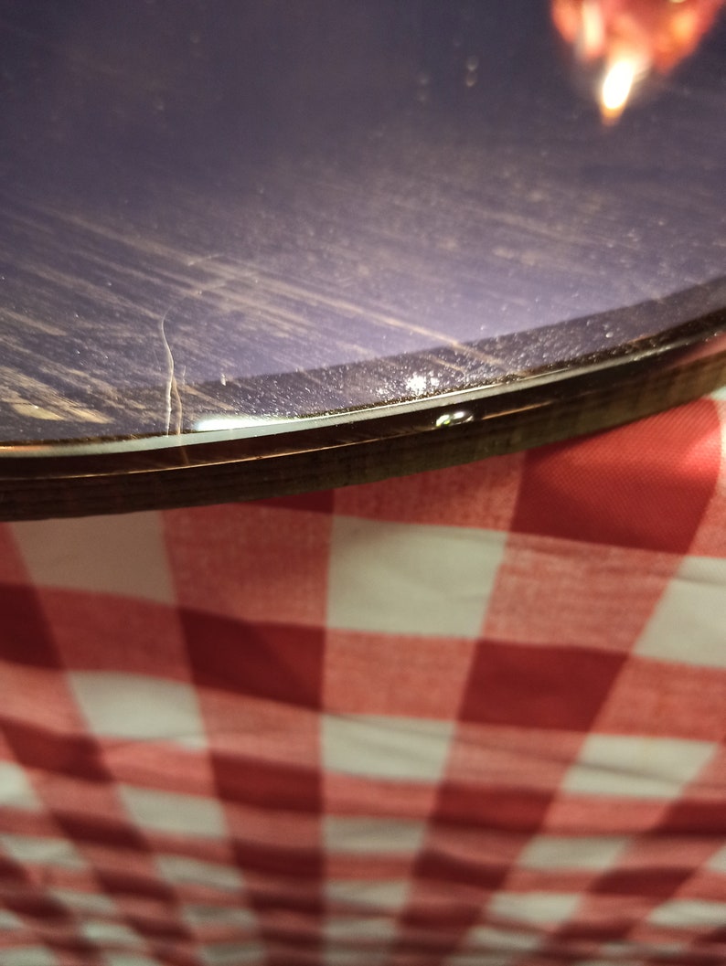 May include: Close-up of the edge of a round, dark purple table with a clear epoxy resin finish. The table is sitting on a red and white checkered tablecloth.