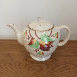 May include: Cream-colored ceramic teapot with a lid and handle. The teapot is decorated with a floral design featuring green, red, and yellow leaves. The teapot is sitting on a wooden surface.