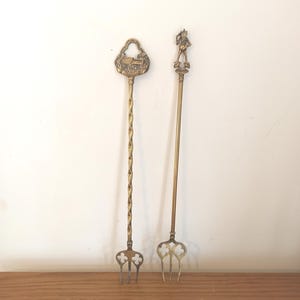 Könnte beinhalten: Zwei antike Serviergabeln aus Messing. Eine Gabel hat einen gedrehten Griff und eine dekorative Oberseite mit einem Gebäudedesign. Die andere Gabel hat einen geraden Griff und eine Figur oben. Beide Gabeln haben ein dreizackiges Design.