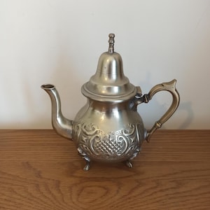 Puede incluir: Tetera plateada con un diseño decorativo floral y de uvas. La tetera tiene un pico curvo, un asa con un acento dorado y una tapa pequeña y ornamentada. Se apoya sobre tres patas pequeñas y se exhibe sobre una superficie de madera.