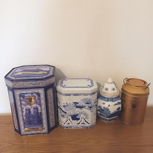 May include: Four decorative storage containers on a wooden surface. Includes a blue and gold tin with a cathedral image, a white and blue porcelain container with a landscape design, a small blue and white jar, and a gold tin.