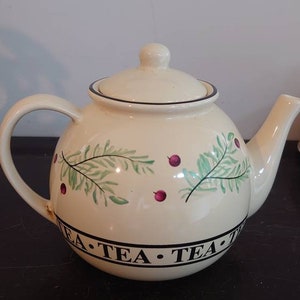 Könnte beinhalten: Eine cremefarbene Teekanne mit grünem und rotem Blumendesign. Die Teekanne hat ein schwarzes Band um den Boden mit den Worten "TEA TEA" darauf.