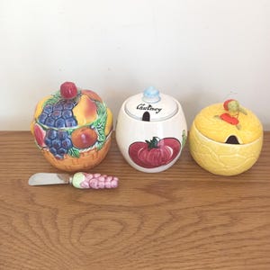 May include: Three ceramic serving dishes with lids, one shaped like a basket of fruit, another with a tomato design and the word "Chutney", and a third with a yellow textured surface. A small knife with a grape-shaped handle is also present.