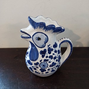 Puede incluir: Una jarra de cerámica blanca con forma de gallo con diseños florales azules y blancos. La jarra tiene un pico y una asa.