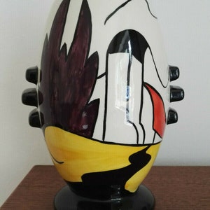 Könnte beinhalten: Eine Keramikvase mit einem Cartoon-artigen Design in Schwarz, Weiß, Rot, Gelb und Lila. Die Vase hat einen runden Körper und einen schwarzen Sockel.