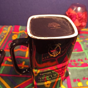 May include: Black ceramic mug with a gold design of a man in a kilt holding a bottle of whiskey. The mug has the words "Johnnie Walker Black Label" and "Old Scotch Whisky" printed on it.