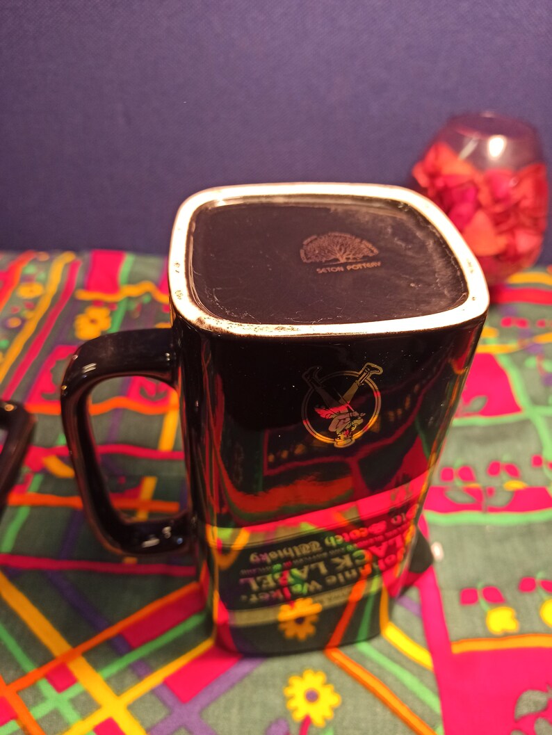 May include: Black ceramic mug with a gold design of a man in a kilt holding a bottle of whiskey. The mug has the words "Johnnie Walker Black Label" and "Old Scotch Whisky" printed on it.