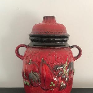 May include: Red ceramic jar with a lid, featuring a black rim and a design of pears and other fruit. The jar has the word "Rumtopf" written on it.