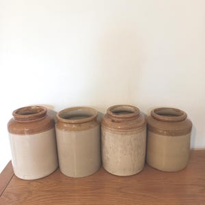 Op de afbeelding: Vier antieke steengoed potten met bruine en beige kleuren. De potten zijn cilindrisch met een brede opening en een afgeronde rand. Ze staan in een rij op een houten oppervlak, met een witte muur op de achtergrond.