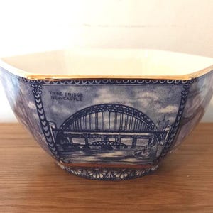May include: A blue and white ceramic bowl with an octagonal shape. The bowl features a detailed illustration of the Tyne Bridge in Newcastle. The rim has a gold trim. The text "Tyne Bridge Newcastle" is visible on the side of the bowl.