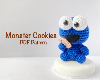 Op de afbeelding: Blauw gehaakt monster cookie amigurumi speelgoed dat een bruine koek vasthoudt. Het speelgoed heeft witte ogen met zwarte pupillen en een zwarte mond. De tekst "Monster Cookies PDF Pattern" staat in bruine tekst onder het speelgoed.