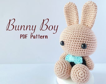 May include: A crocheted bunny toy in a light brown color, with black eyes and a light blue bow tie. The text "Bunny Boy PDF Pattern" is written in a cursive font. The bunny is sitting on a white base.