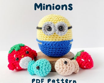Peut inclure: Divers personnages Minions et fraises crochetés. Le Minion central est jaune et bleu, avec de grands yeux gris et des lunettes noires. Deux fraises rouges et deux petites figurines en forme de poisson sont également présentes. Le texte "Minions" et "PDF Pattern" sont affichés.
