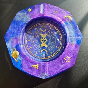 Könnte beinhalten: Ein Aschenbecher aus lila und blauem Harz mit einem goldenen Himmelsdesign. Das Design zeigt eine Sonne, einen Mond und Sterne.
