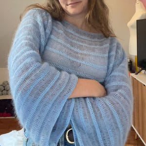 Puede incluir: Un suéter azul claro de manga larga con un tejido suelto y abierto. El suéter tiene cuello barco y mangas acampanadas. La prenda está hecha de un material suave, posiblemente tejido. El suéter se usa con jeans azules.