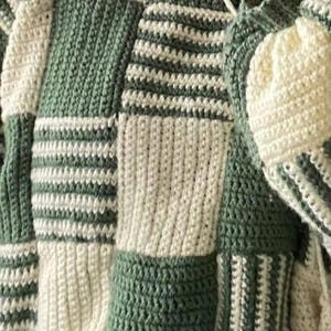 Peut inclure: Un pull crocheté vert et blanc avec un motif à carreaux. Le pull a des manches longues et une coupe ample.