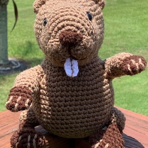 May include: A brown crocheted beaver plush toy with white teeth and brown accents on the paws. The toy is sitting on a wooden surface.