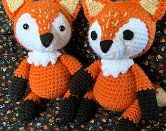 May include: Two handmade crochet fox dolls. The foxes have orange bodies, white faces, and black paws. They have tan ears and white ruffs around their necks. The dolls are sitting on a patterned fabric background.