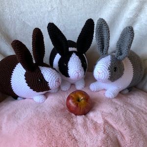 May include: Three crocheted stuffed bunny rabbits in black and white, brown and white, and gray and white, sitting on a pink fuzzy surface with a red apple in front of them.