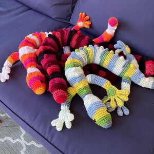 May include: Several colorful, crocheted lizard-like toys are arranged on a purple surface. The toys feature vibrant stripes of red, orange, pink, yellow, blue, and green. Each has a long, segmented body, small legs, and a head with a small, round eye.