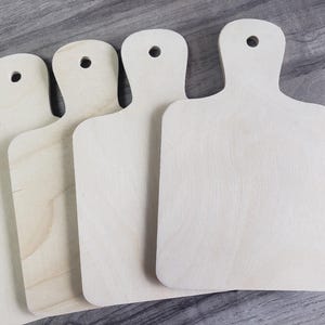 May include: A collection of unfinished wooden cutting boards with handles. The boards are rectangular with rounded corners and a hole in the handle for hanging. The wood has a light, natural color and visible grain.