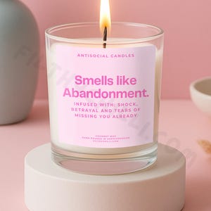 May include: A lit candle in a clear glass jar with a pink label that reads "Smells like Abandonment." The candle is on a white circular stand. The background is a soft pink.