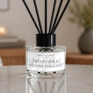 May include: A clear glass reed diffuser bottle with a black cap and six black reeds. The white rectangular label features the text "TWENTY-ONE & FUCKING FABULOUS" in black capital letters, with smaller text above reading "Intimate Scent Collection".