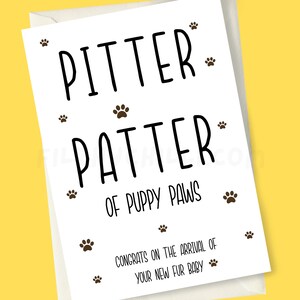 May include: A white card with black text that reads "Pitter Patter of Puppy Paws" and "Congrats on the arrival of your new fur baby". The card is decorated with brown paw prints.