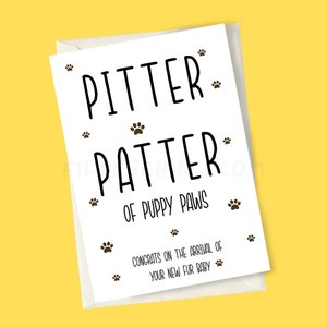 May include: White card with black text that reads "Pitter Patter of Puppy Paws" with paw prints scattered around the text. The card also has the text "Congrats on the arrival of your new fur baby" with paw prints scattered around the text.