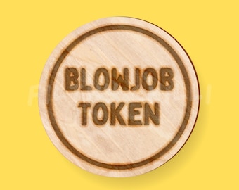 May include: A round wooden token with the words "BLOWJOB TOKEN" etched into the surface. The token has a light brown color with a darker brown border and text. The background is a solid yellow.