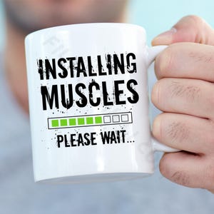 May include: White ceramic coffee mug with black text that reads "Installing Muscles" and a progress bar with green filled bars and empty white bars. The text "Please Wait..." is below the progress bar.