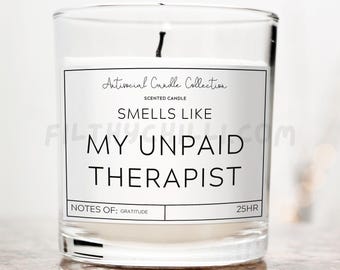 Op de afbeelding: Een glazen kaars pot met een wit label waarop staat "Antisocial Candle Collection" en "Smells Like My Unpaid Therapist". Het label bevat ook "Notes of: Gratitude" en "25HR".