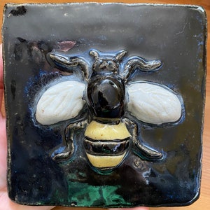 May include: A ceramic tile featuring a three-dimensional bee sculpture. The bee is black and yellow with white wings and is set against a dark blue background.
