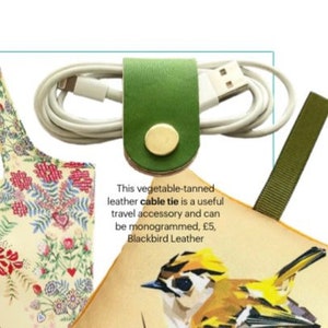 May include: Green leather cable tie with a brass button. The cable tie is wrapped around a white charging cable. The text "This vegetable-tanned leather cable tie is a useful travel accessory and can be monogrammed, &pound;5, Blackbird Leather" is visible below the cable tie.