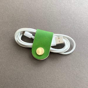 May include: A green leather cord organiser with a brass button. The organiser is holding a white charging cable.