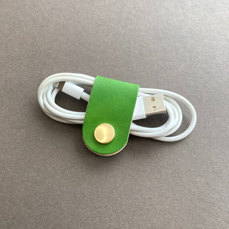 May include: A green leather cord organiser with a brass button. The organiser is holding a white charging cable.
