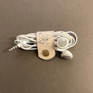 May include: A white leather cord organiser with a speckled pattern and a brass button. The organiser is holding a pair of white earbuds.