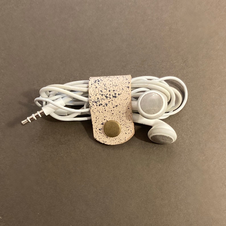 May include: A white leather cord organiser with a speckled pattern and a brass button. The organiser is holding a pair of white earbuds.