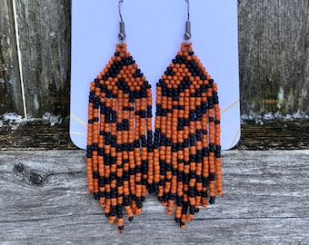 May include: A pair of orange and black beaded dangle earrings with silver hooks. The earrings are displayed on a white card with the text "J+G" printed on it.