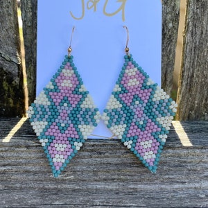 Pode incluir: Um par de brincos em forma de diamante com um padrão geométrico de contas rosa, brancas e azul-turquesa. Os brincos estão em um cartão branco com o texto "J+G" impresso nele.
