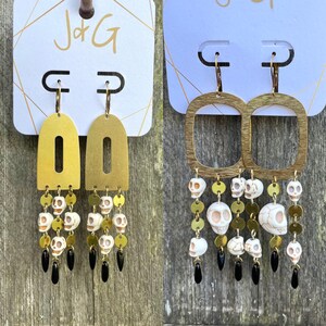 Könnte beinhalten: Ein Paar goldene Ohrringe mit Totenkopf- und Knochenmotiv. Die Ohrringe sind aus Messing und haben eine baumelnde Kette mit weißen Totenkopfperlen. Die Ohrringe befinden sich auf einer weißen Karte mit dem aufgedruckten Text "J+G".